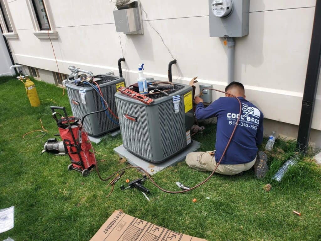 A technician in a blue shirt is kneeling on the grass, diligently performing AC maintenance beside a building. Flanked by two outdoor air conditioning units and an array of tools, he demonstrates precision and expertise in AC installation & replacement in Nassau County.