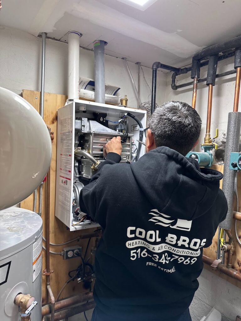 A technician in a black hoodie with "Cool Bros Heating & Air Conditioning" is repairing a boiler in a utility room. Surrounded by exposed pipes and a water heater, he expertly handles both the current fix and any AC repair needs for Nassau County residents.