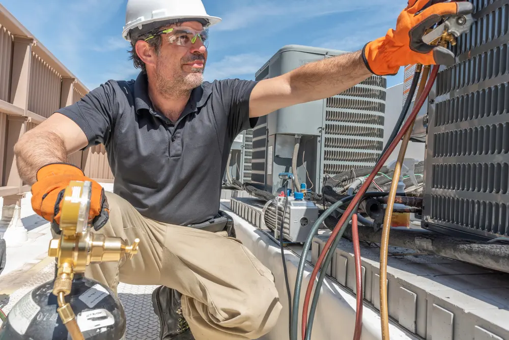 A technician in a hard hat and safety gloves is working on an air conditioning unit on a rooftop, performing AC repair in Nassau County. He's checking equipment with tools, surrounded by various HVAC components under a clear blue sky.