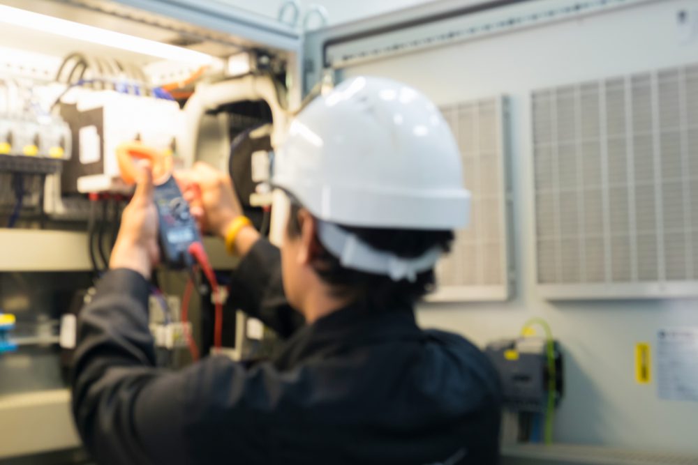 An electrician in a white hard hat uses a multimeter to check electrical equipment inside an industrial control panel, ensuring top-notch AC maintenance in Nassau County. The setting appears to be a maintenance or inspection site.