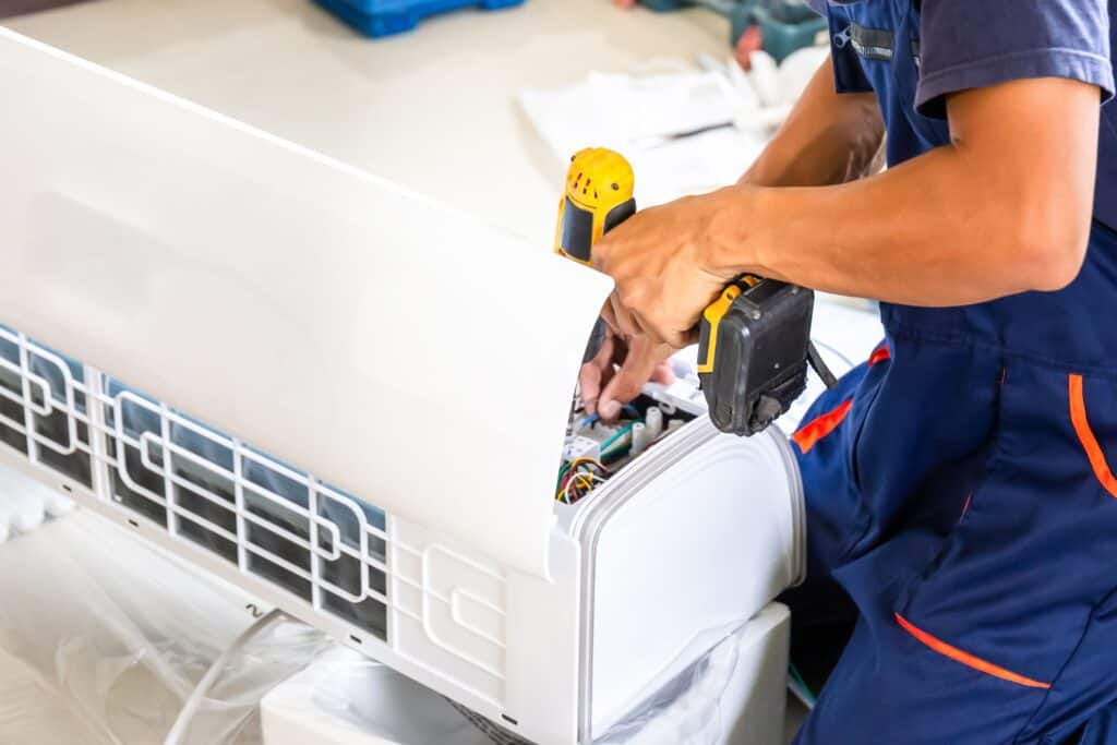 A person in uniform is expertly installing or repairing a wall-mounted air conditioning unit. Using a cordless drill among other tools, they focus on the internal components of the unit. The indoor, well-lit setting signals a professional AC Installation & Replacement Nassau County service at work.