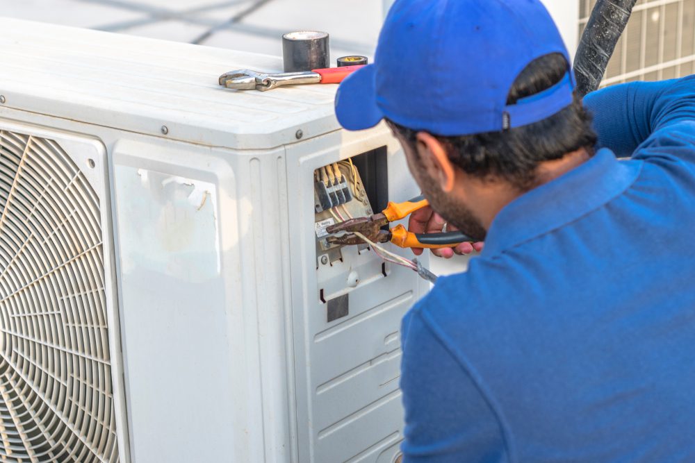 A technician in a blue uniform and cap carefully works on an outdoor air conditioning unit on a rooftop in Nassau County NY, using pliers to adjust wires. Nearby, a wrench and a screwdriver rest atop the unit, embodying expert conditioning service in this bustling area.