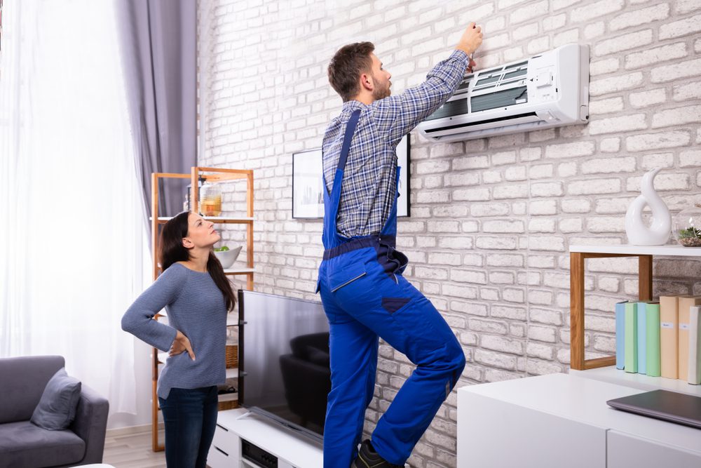 A technician in blue overalls stands on a stepladder, expertly conducting AC maintenance Nassau County style, while a woman watches. The room features a brick wall, TV, shelves, and gray curtains.