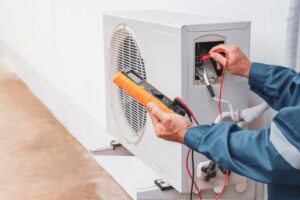 A technician in a blue jacket performs AC maintenance in Nassau County, using a multimeter to test electrical components of an outdoor air conditioning unit on a concrete pad. The technician expertly holds red and black probes at the unit's panel, ensuring optimal performance.