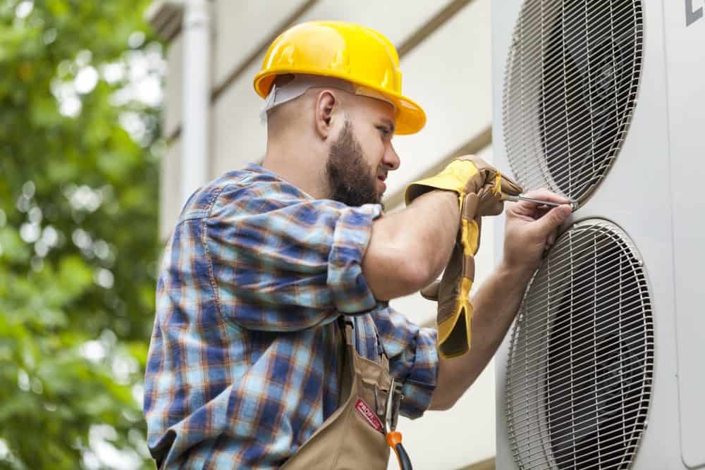 A worker in a yellow hard hat and plaid shirt expertly repairs an outdoor air conditioning unit with a screwdriver, set against lush green foliage and a beige building, showcasing the precision of AC Installation & Replacement Nassau County.