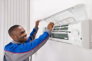 A technician smiles as he inspects and repairs an air conditioning unit, holding the cover open. He is wearing a gray and blue uniform. The background features white blinds and a white wall.