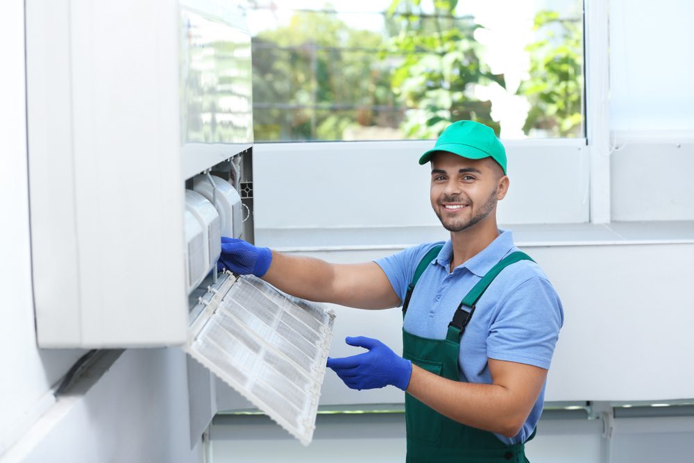 A technician wearing a green cap, blue gloves, and overalls is smiling while cleaning an air conditioner filter indoors. He stands near a window with a blurred view of greenery outside.