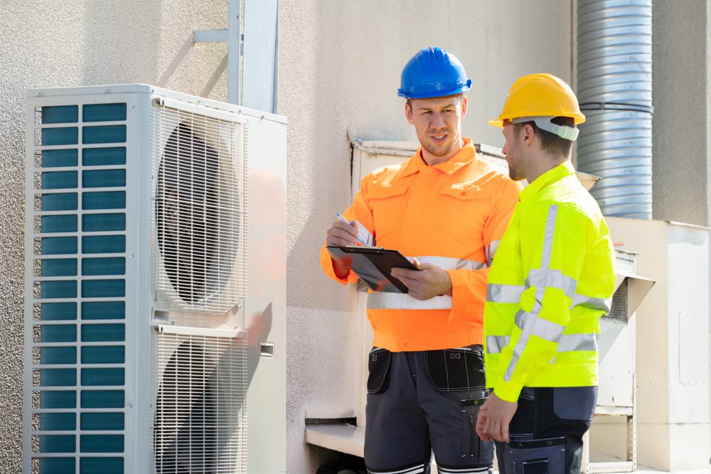 Two construction workers in high-visibility jackets and helmets inspect an outdoor air conditioning unit. One holds a clipboard, taking notes, while they discuss the unit's condition.