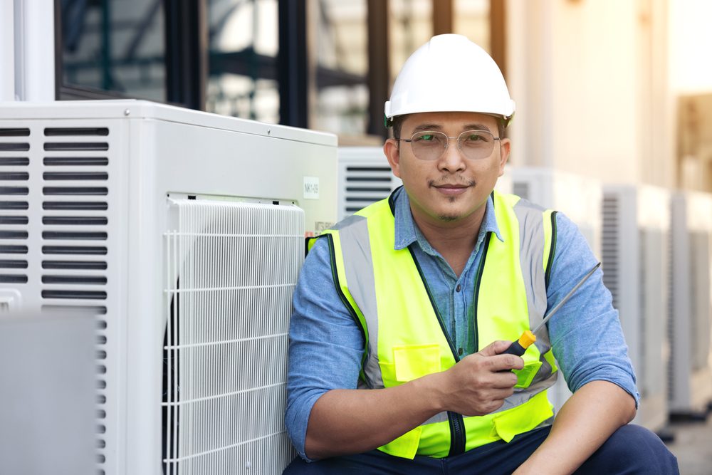 A man wearing a white hard hat and a yellow safety vest sits next to an outdoor air conditioning unit. He is holding a walkie-talkie and looking at the camera. The background features more air conditioning units.