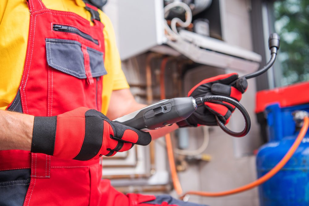 A technician in red overalls and gloves holds a handheld device with a flexible nozzle, likely inspecting for gas leaks. In the background, industrial equipment and blue gas cylinders are visible.