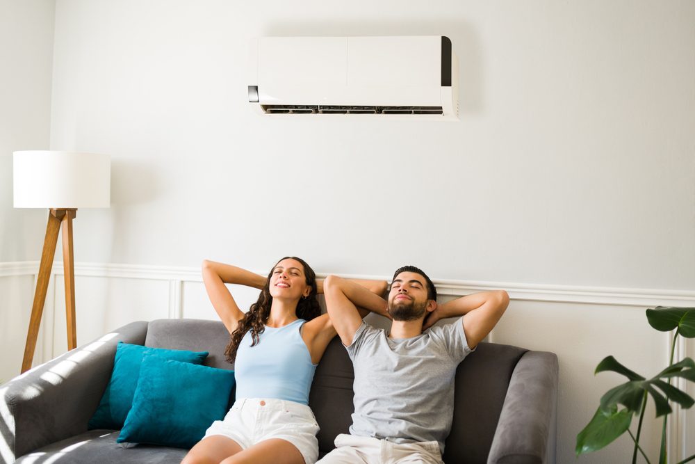 A man and woman relax on a gray sofa with their arms behind their heads, sitting under a wall-mounted air conditioner. The room is bright, with a floor lamp and a potted plant visible in the corner.