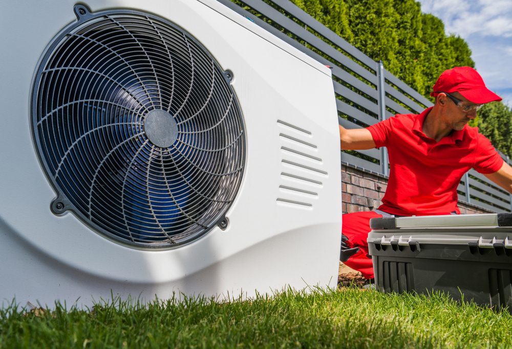 A technician in a red uniform sits on the grass next to an outdoor air conditioning unit, inspecting it. A toolbox is open nearby. The background includes a metal fence and green trees.