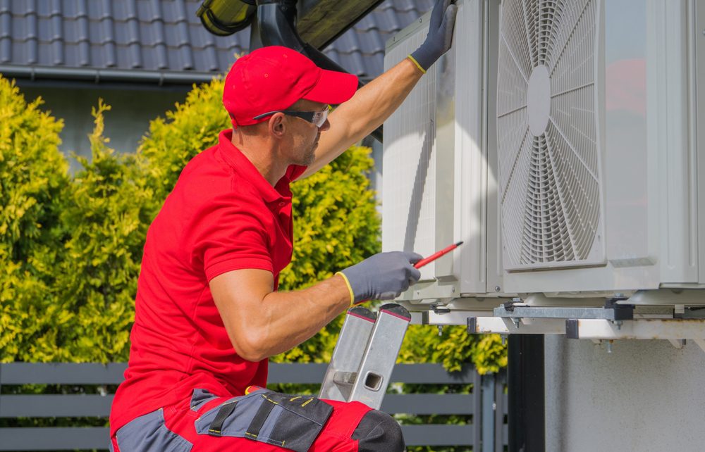 A technician in a red uniform and cap is working on an outdoor air conditioning unit. He uses tools to adjust components while standing on a ladder. Green bushes and a gray fence are in the background, along with the corner of a building.