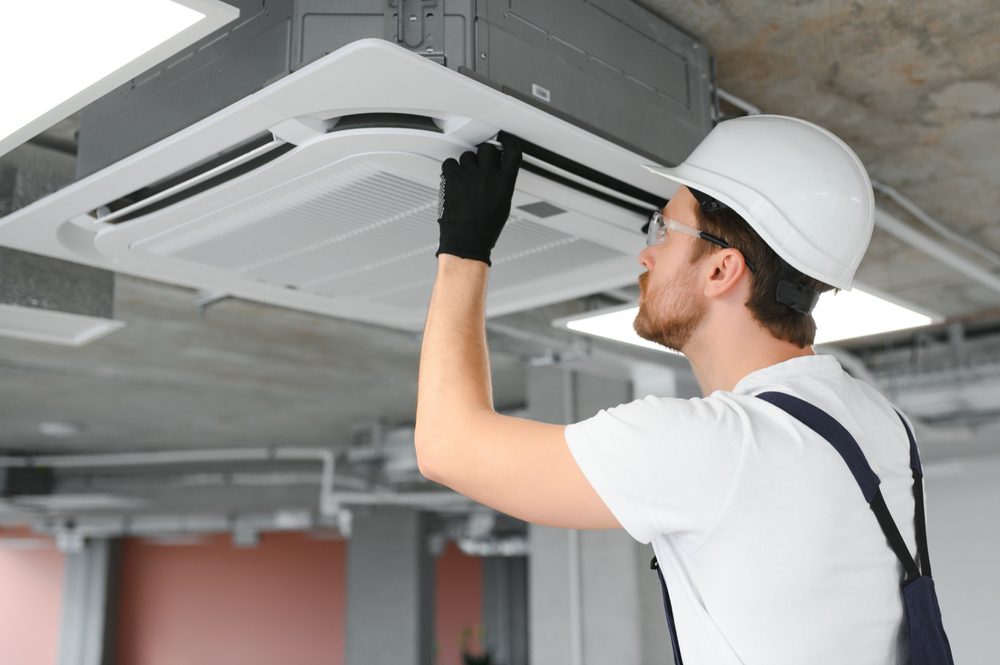 A technician wearing a hard hat and gloves is inspecting a ceiling-mounted air conditioning unit. He is looking closely at the unit in an industrial or commercial setting. The ceiling is made of exposed concrete.