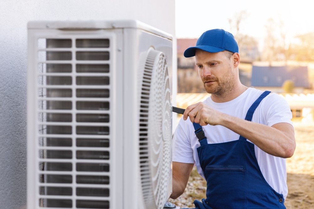 A technician in blue overalls and a cap adjusts an outdoor air conditioning unit with a screwdriver. The background shows a sunny day and a blurred view of trees and rooftops.