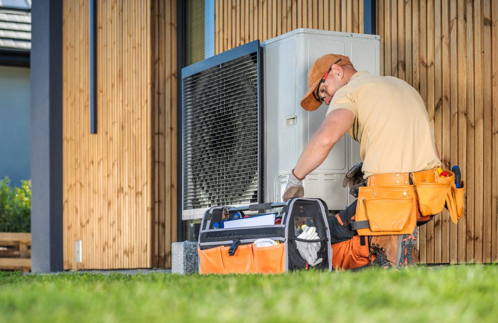 A technician in an orange cap and tool belt works on an outdoor air conditioning unit, surrounded by a toolbox and equipment. The setting is a wooden exterior of a house, with grass in the foreground.