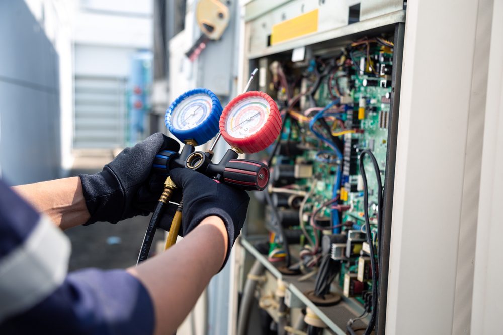 A person wearing gloves uses a manifold gauge set with blue and red dials to inspect or service an open electrical panel, containing numerous wires and circuit boards, outdoors.