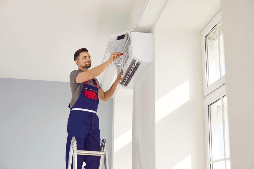 A technician in a uniform stands on a ladder, installing or repairing an air conditioning unit on a wall. He holds a tool and appears focused on his work. Sunlight streams in through large windows, illuminating the bright room.