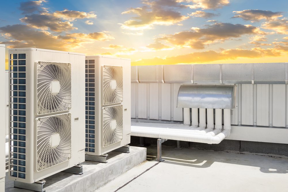 Air conditioning units on a rooftop with a scenic sunset in the background. The sky features clouds and a warm glow, creating a contrast between the industrial equipment and the natural beauty of the sunset.