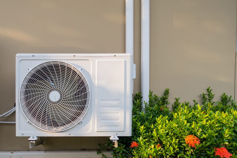 A white outdoor air conditioning unit is mounted on a beige wall. Lush green shrubs with small orange flowers are growing beneath the unit, adding a touch of nature to the scene.