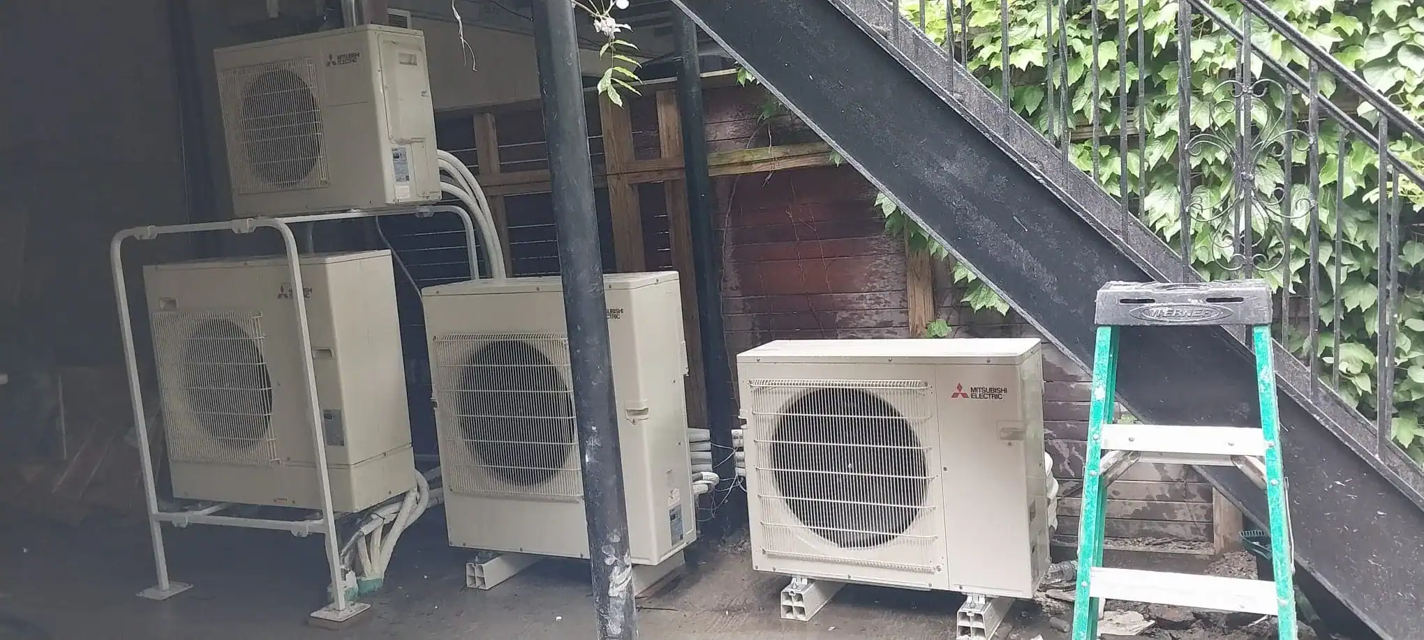 Four outdoor air conditioning units, installed by an HVAC Contractor Nassau County, are set beneath a metal staircase. A green step ladder stands nearby, with a wooden fence and leafy vines in the NY background.