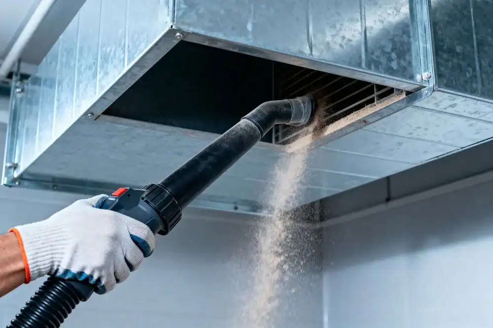 A person wearing a glove uses a vacuum hose to clean dust from an air duct vent in a building ceiling—typical work for an HVAC Contractor in Nassau County, NY.