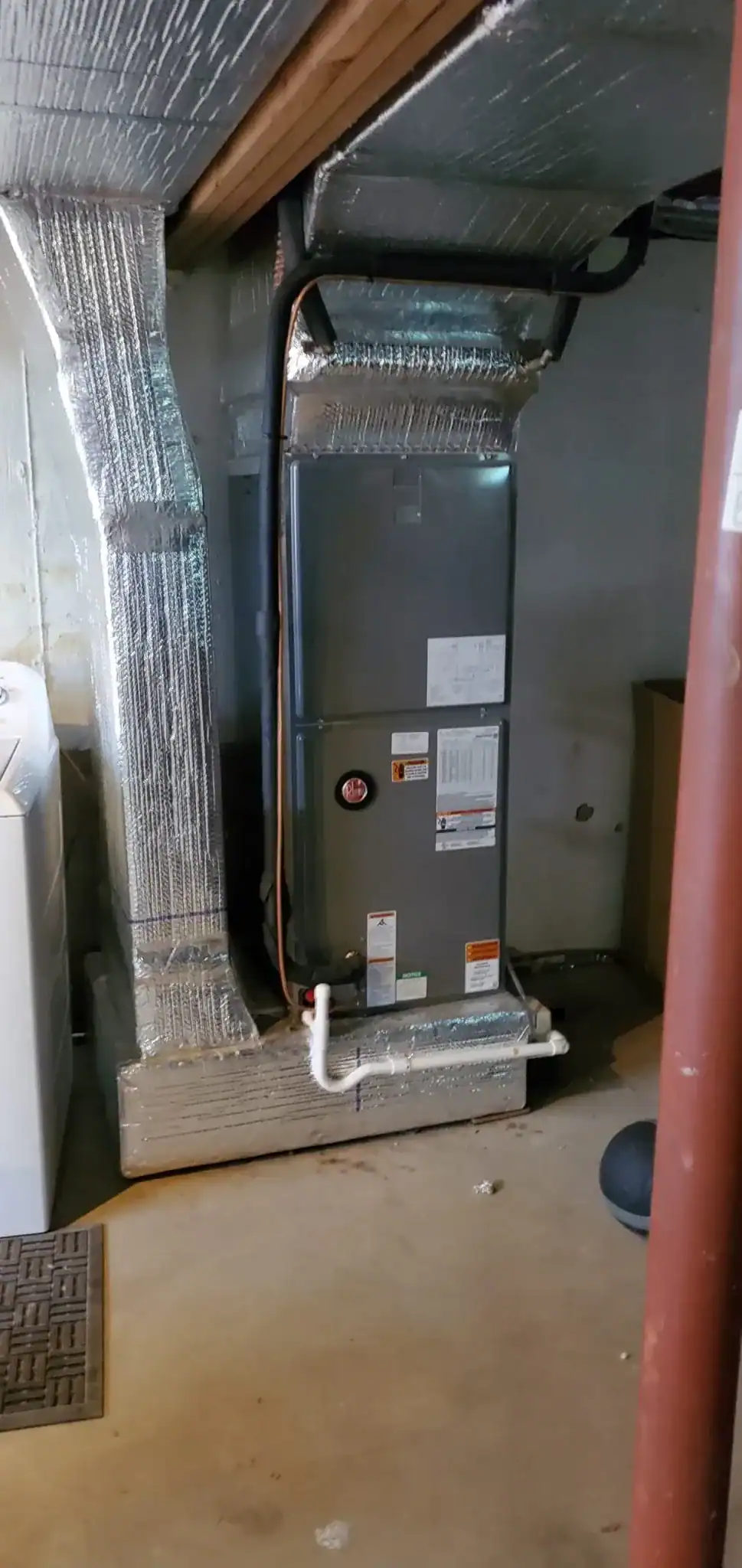 A central HVAC furnace unit with insulated ductwork is installed in a NY basement by an expert HVAC Contractor Nassau County. Nearby, a washing machine is partially visible on the left, and a brown box and support post are on the right.