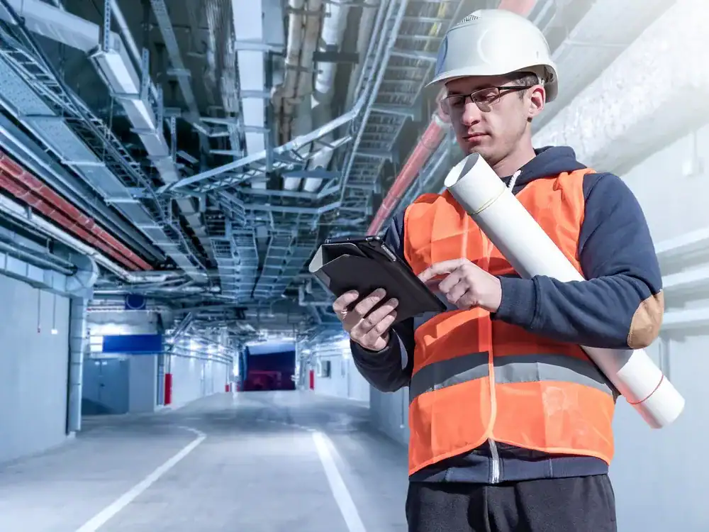 A construction worker wearing a hard hat and orange safety vest stands in an industrial tunnel, holding blueprints and using a tablet&mdash;typical of an HVAC Contractor Nassau County, NY&mdash;surrounded by exposed pipes and overhead wiring.