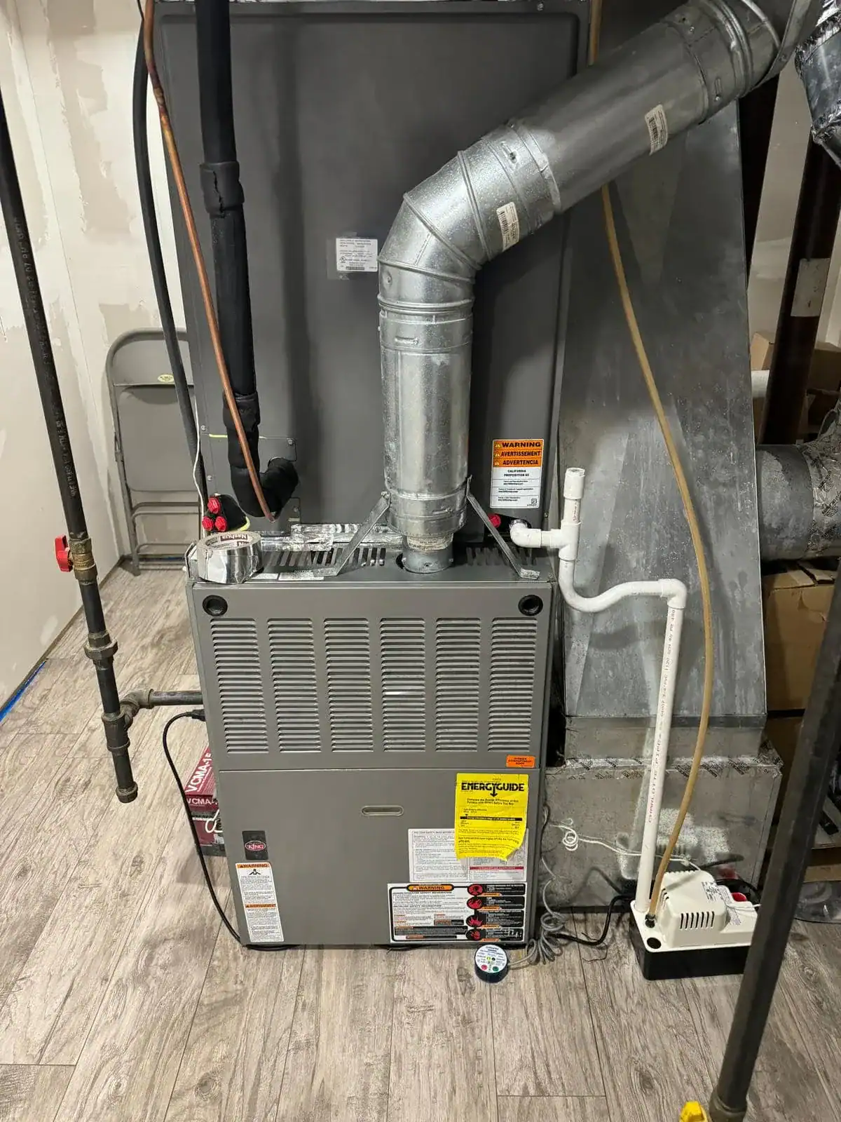 A gray furnace unit with metal ductwork, white PVC pipes, and warning labels is installed on a wood-patterned floor in a utility room. A small white pump sits on the floor next to the unit&mdash;installed by an expert HVAC Contractor Nassau County.