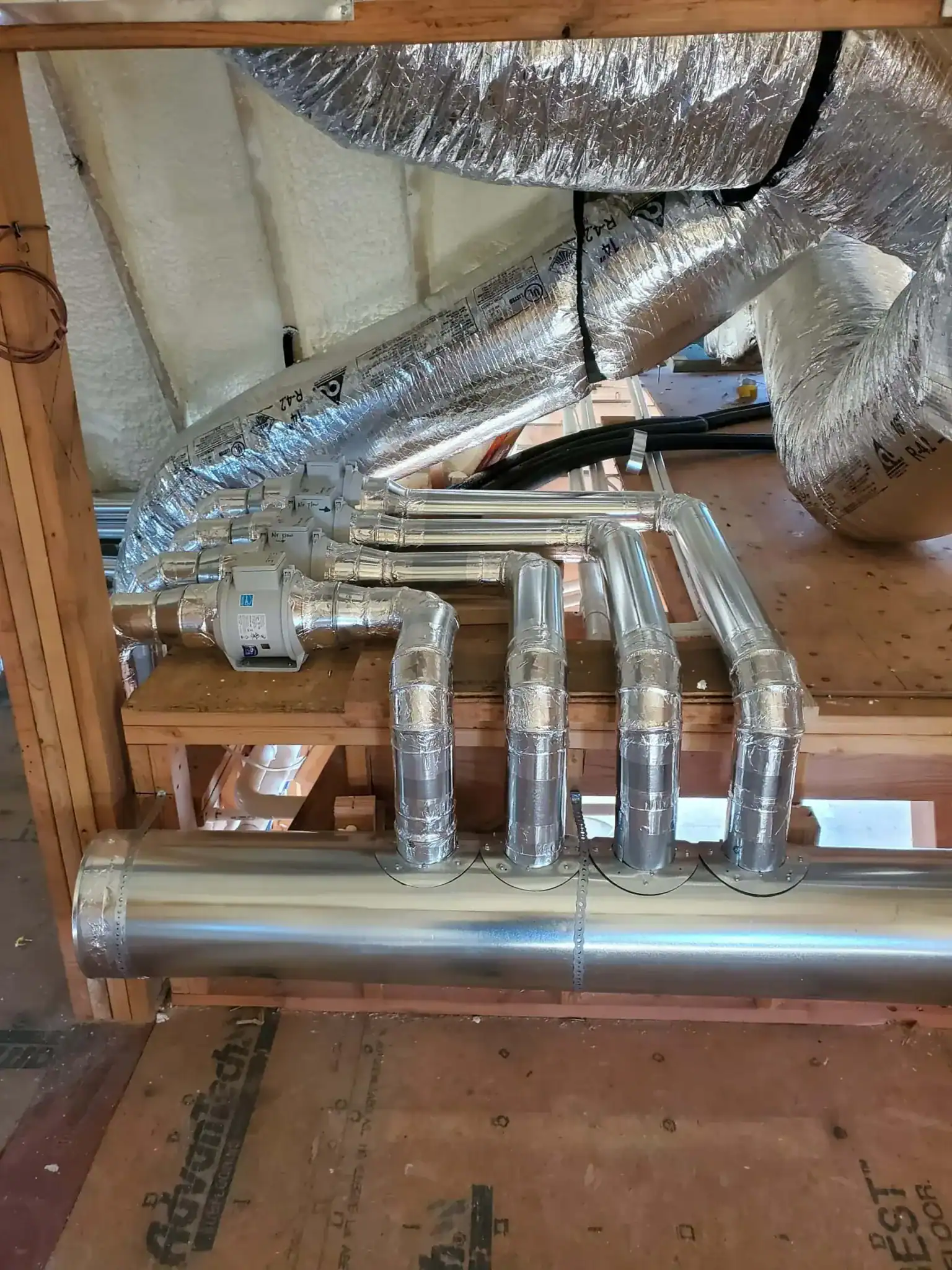 Shiny metal HVAC ducts and pipes run along a wooden structure with insulation on the walls and ceiling. Installed by an expert HVAC Contractor Nassau County, NY, five ducts connect vertically from a large horizontal pipe to vents above.