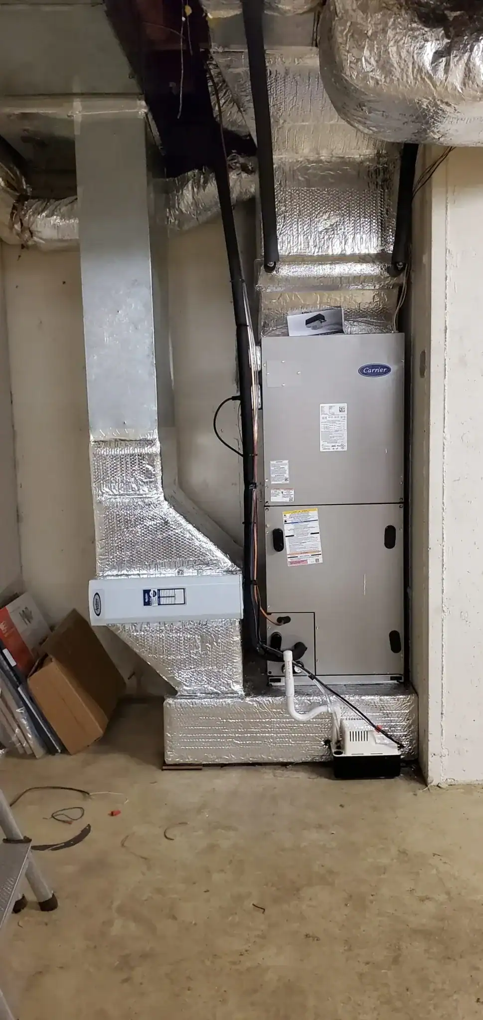 A silver HVAC unit with wrapped ductwork stands against a basement wall. Nearby, there are some cardboard boxes and pipes, with exposed wiring and an air filter compartment visible. Installed by an expert HVAC Contractor Nassau County, NY, the floor is unfinished concrete.