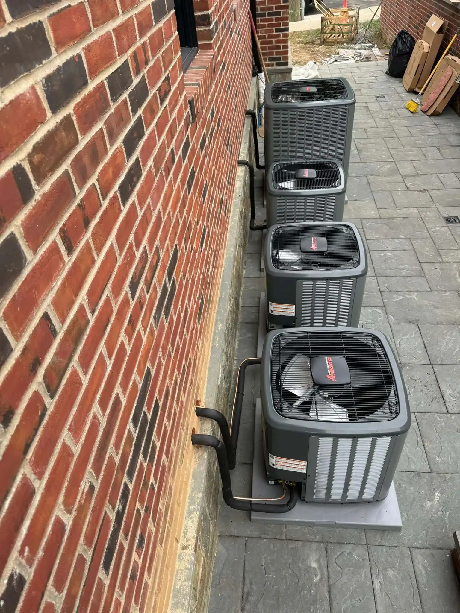Three outdoor air conditioning units are installed in a row along a brick building on a paved patio area by an expert HVAC Contractor Nassau County, NY; black pipes connect the units to the building.
