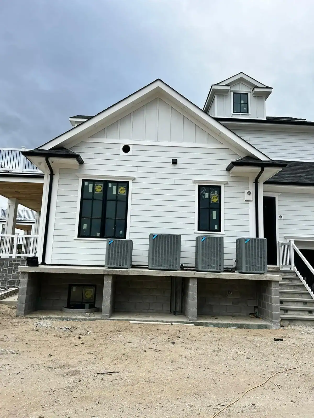 A white house under construction in NY, with three large air conditioning units installed by an HVAC Contractor Nassau County, sits on a raised concrete platform. The ground is unfinished, and several windows remain covered with stickers.