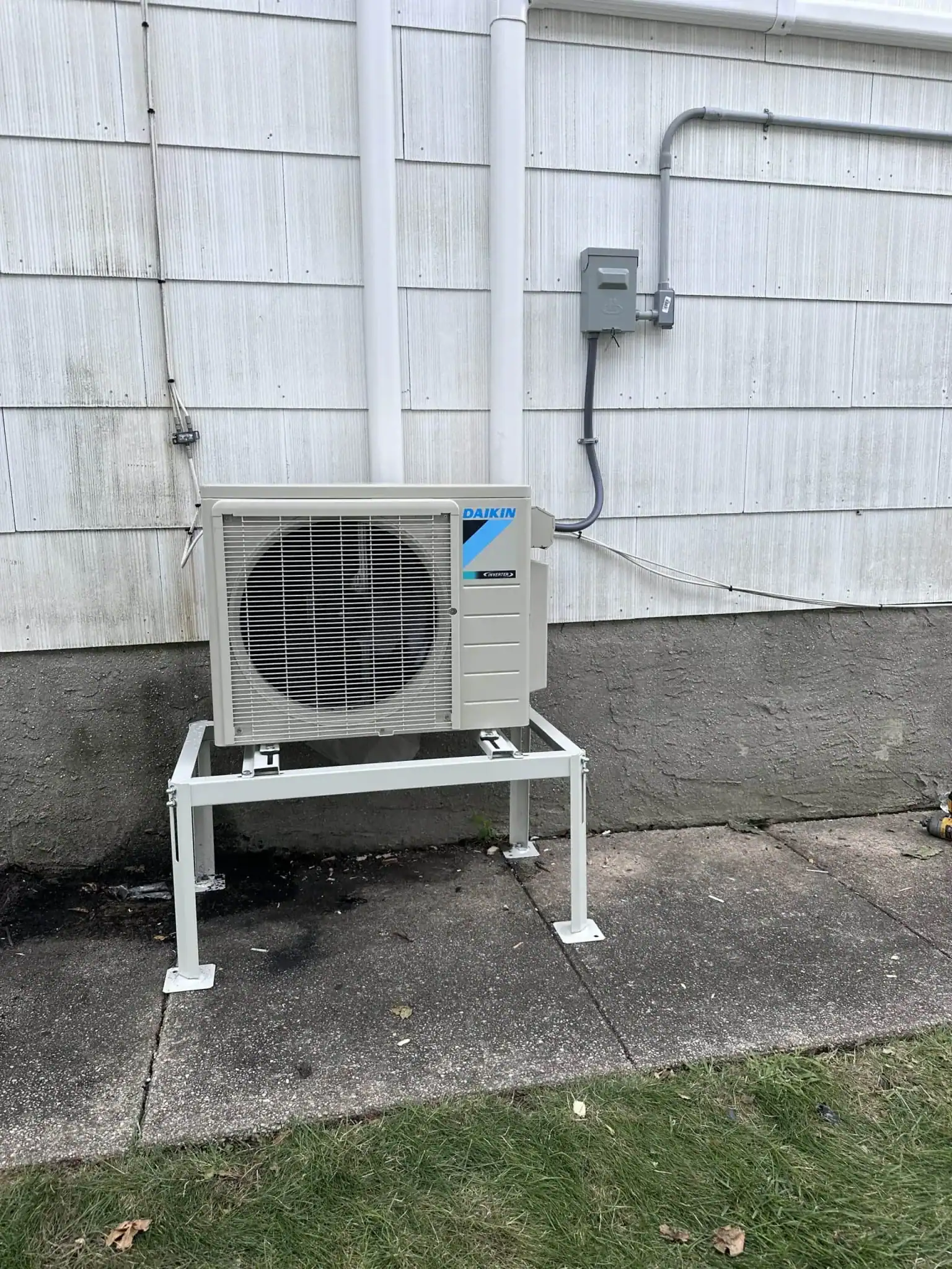 A Daikin outdoor air conditioning unit, installed by an HVAC Contractor Nassau County, NY, is mounted on a metal stand next to a white-sided building above a concrete walkway and beside a grassy lawn.