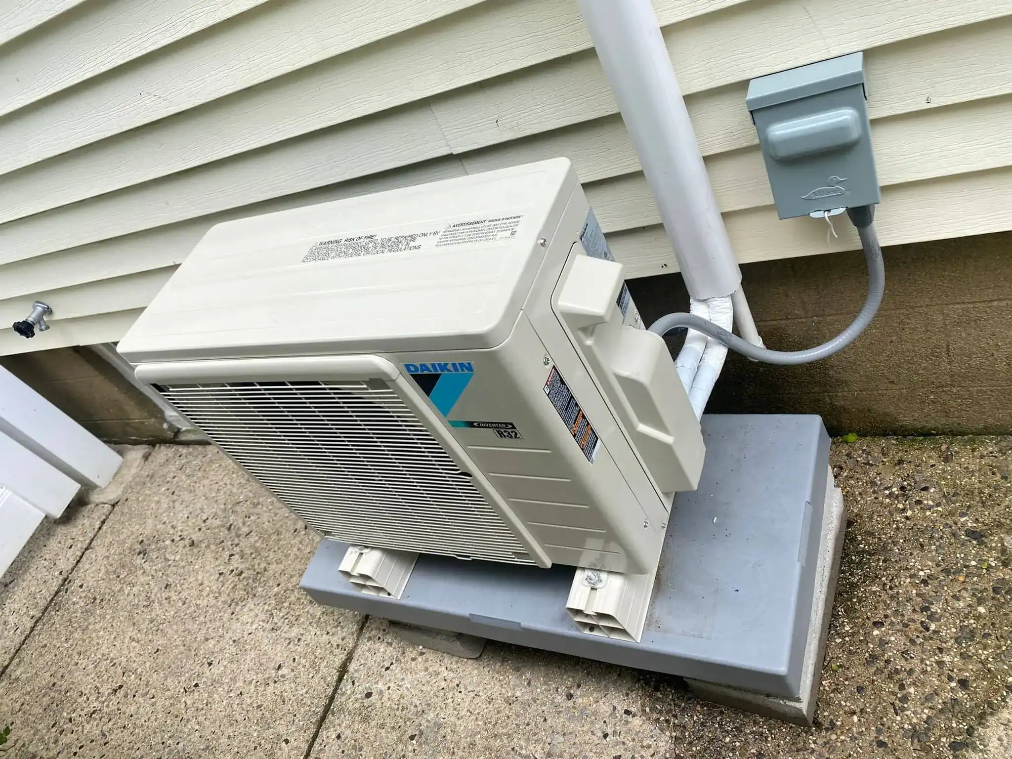 A Daikin outdoor air conditioning unit, installed by an HVAC Contractor Nassau County, NY, is mounted on a raised platform next to a house with light-colored siding and a concrete foundation, with connected power and refrigerant lines.