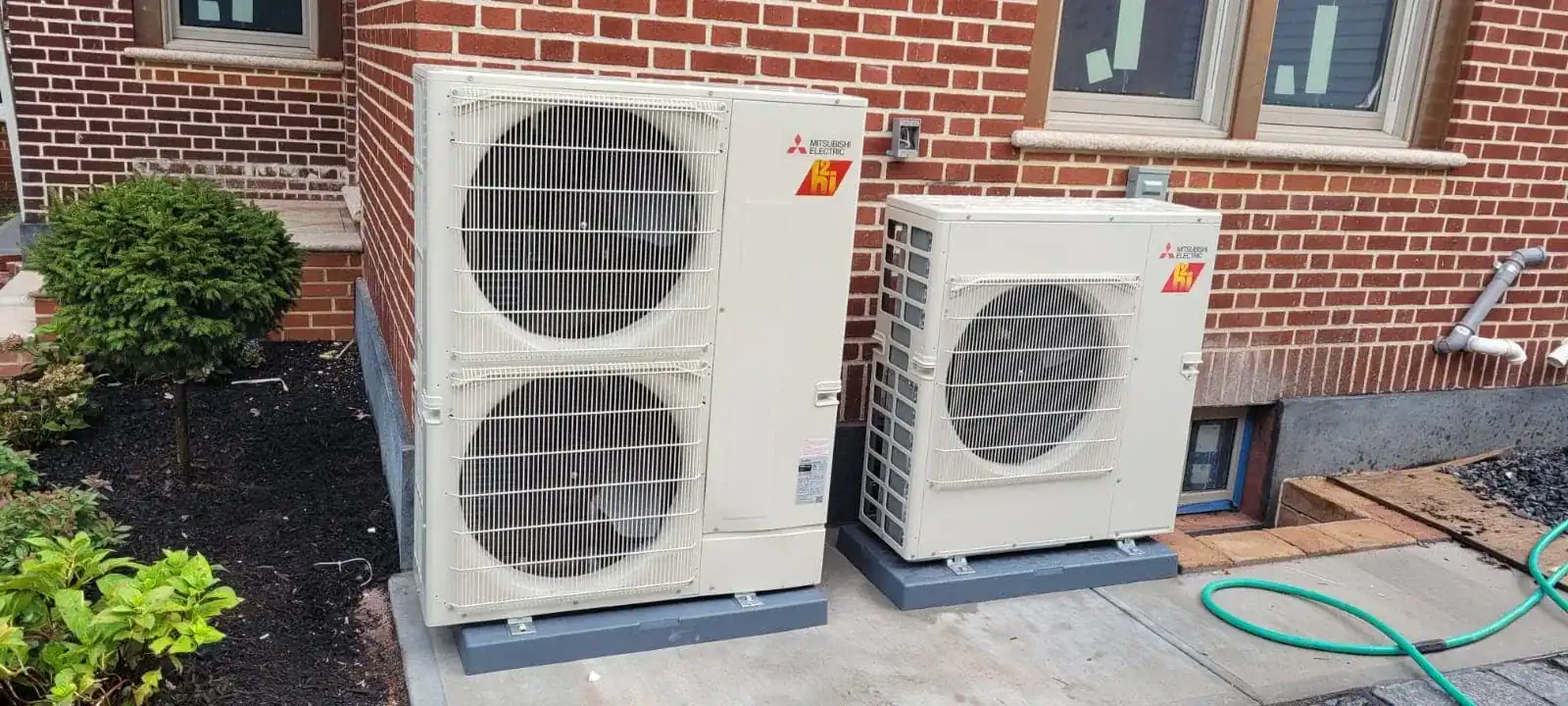 Two large white outdoor air conditioning units sit on concrete pads next to a brick building in NY. A green garden hose lies nearby, and plants with mulch line the left side&mdash;an example of quality work by an HVAC contractor Nassau County trusts.