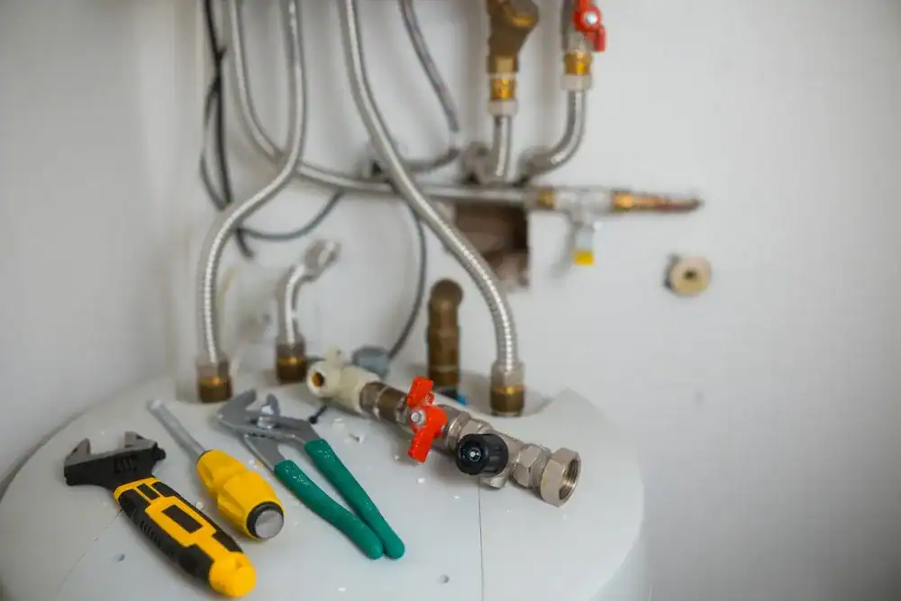 Assorted plumbing tools and a pipe fitting with valves are laid out on top of a white water heater, often seen during inspections by an HVAC Contractor Nassau County, NY, with several metal pipes and connections visible on the wall behind them.