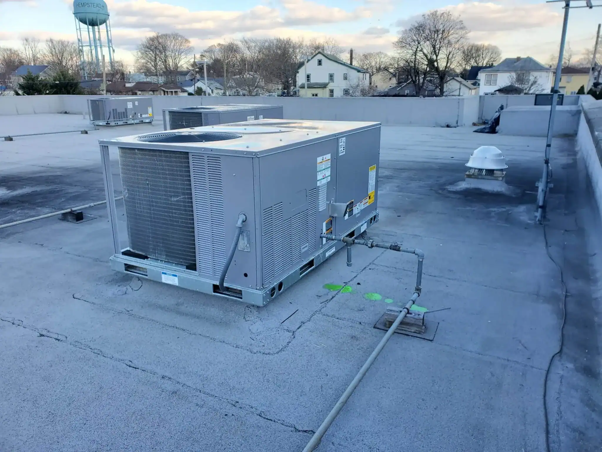 A large HVAC unit, installed by an HVAC Contractor Nassau County, NY, sits on a flat gray concrete rooftop surrounded by pipes and vents. Nearby rooftops, leafless trees, and a water tower appear beneath a partly cloudy sky.