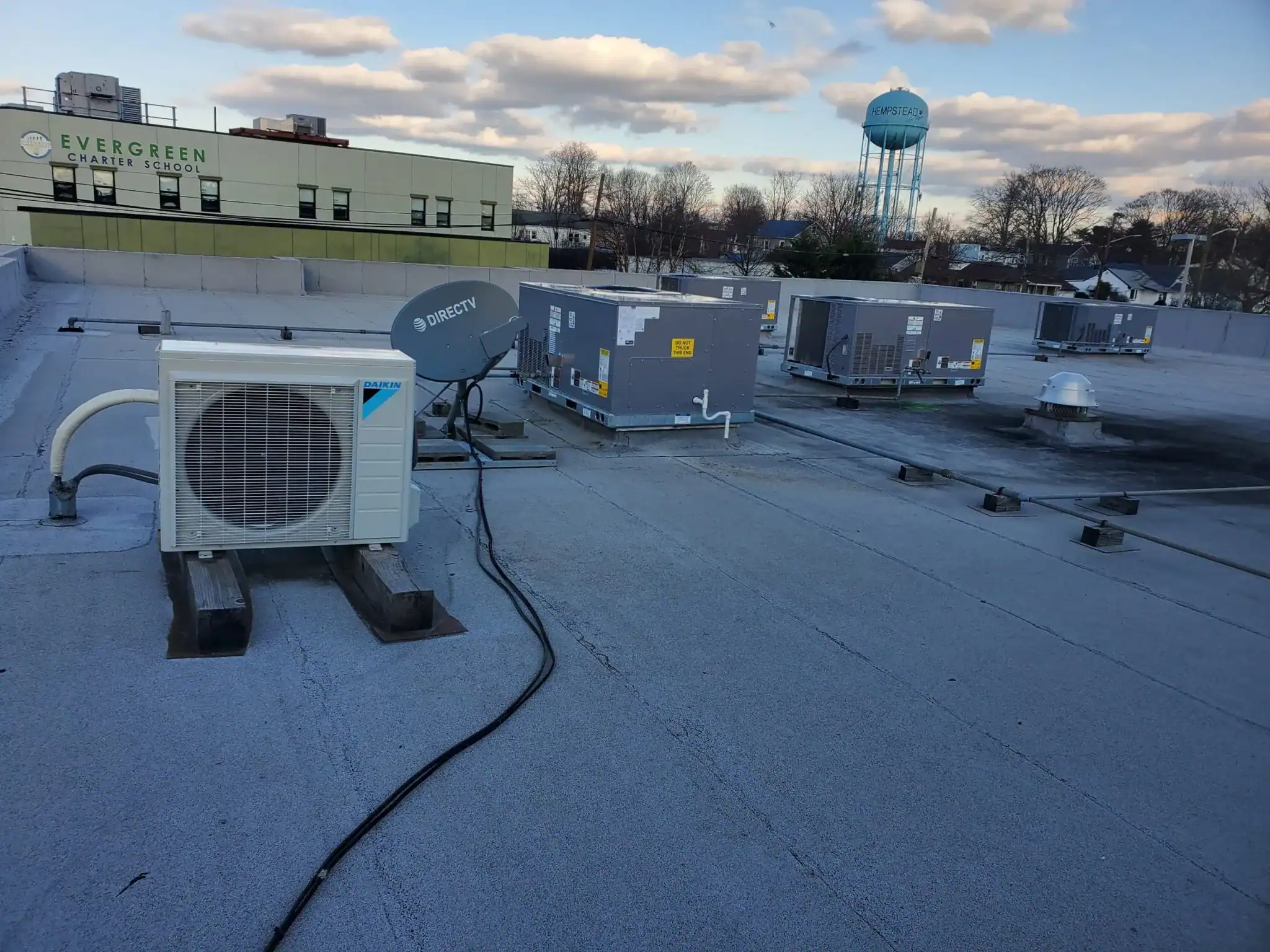 Rooftop view with HVAC units and cables, under a partly cloudy sky. Buildings, bare trees, and a blue "Drexel" water tower are in the background. The "Evergreen Charter School" sign is visible&mdash;ideal for an HVAC Contractor Nassau County, NY project.