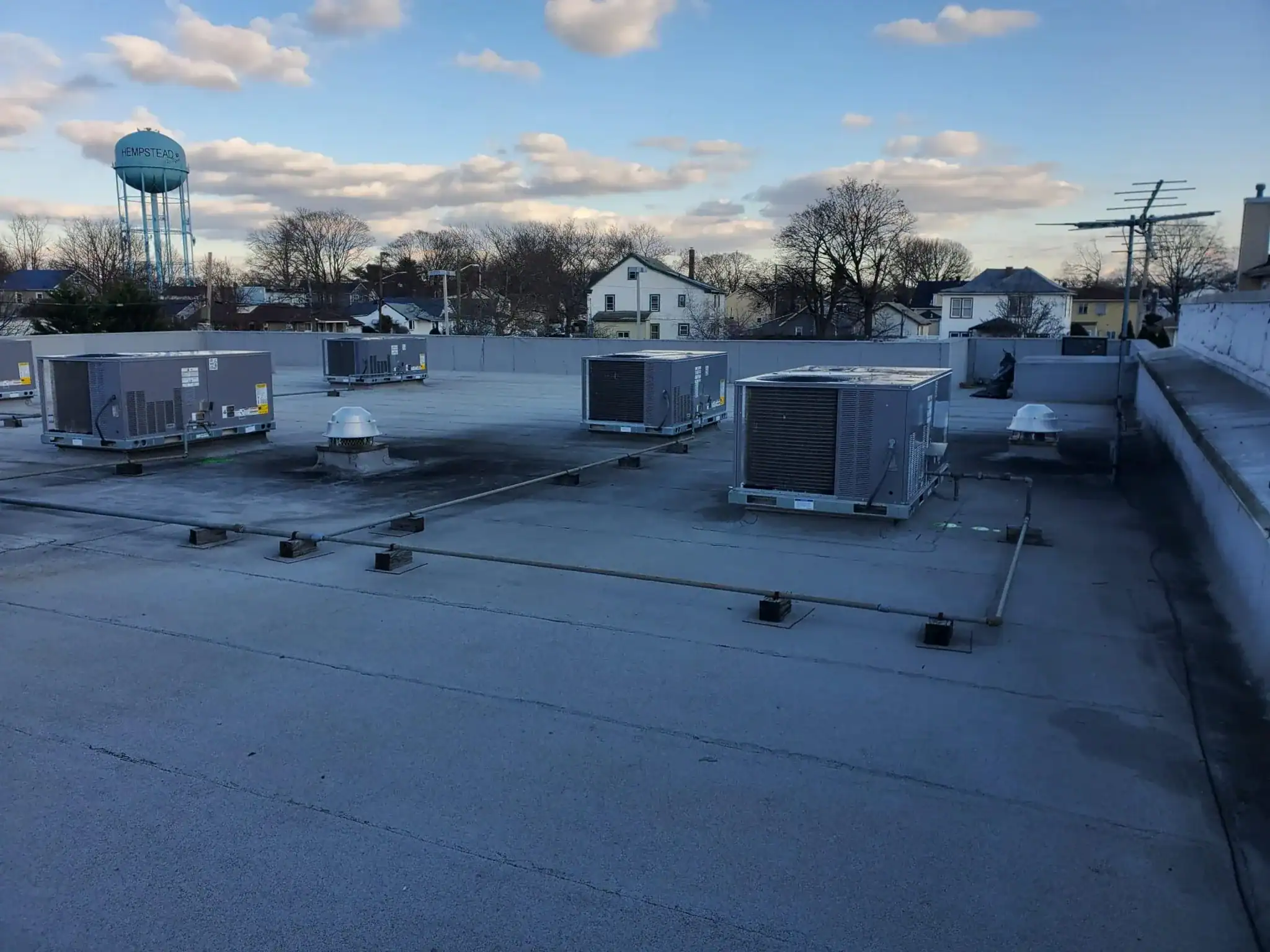 Rooftop view with several HVAC units and ventilation systems under a partly cloudy NY sky; houses, trees, and a water tower are visible in the background&mdash;ideal for any HVAC Contractor Nassau County project.