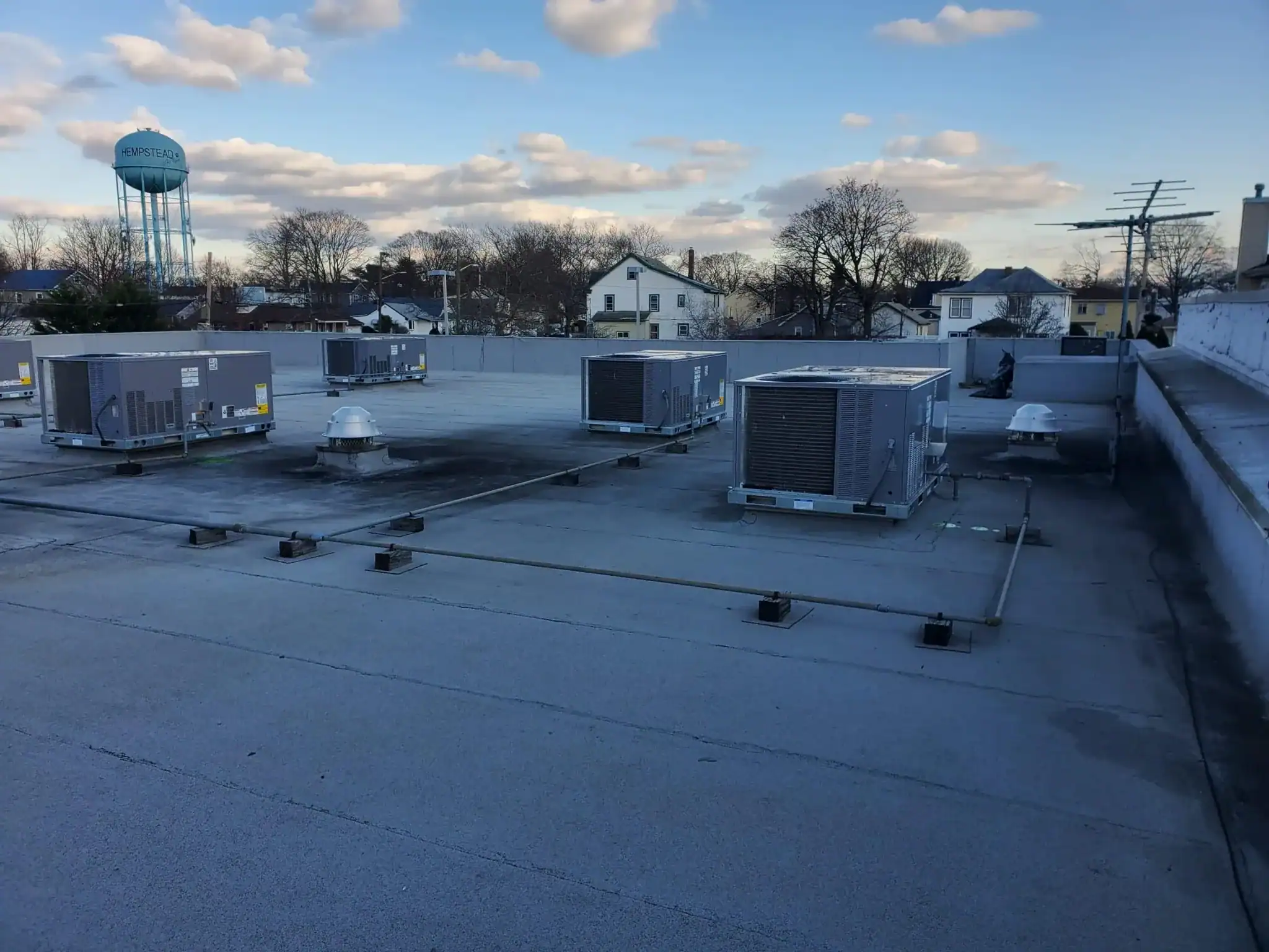 A flat rooftop in NY with several large HVAC units, two vent pipes, and a clear sky with scattered clouds. Trees, houses, and a water tower are visible in the background&mdash;ideal for any HVAC contractor Nassau County project.