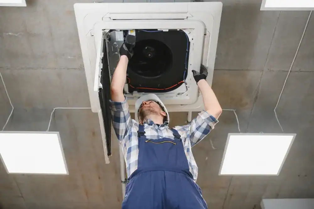 A worker in blue overalls and a hard hat, likely an HVAC contractor in Nassau County, NY, repairs or installs a ceiling-mounted air conditioning unit, standing on the floor and looking up at the open panel.