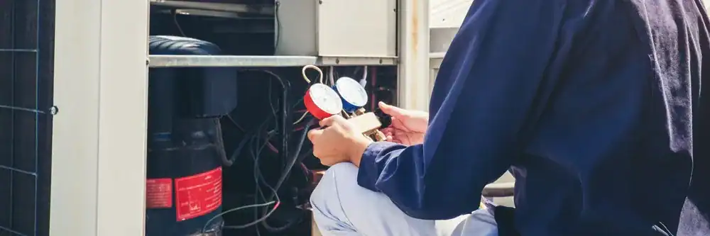 A technician in a blue uniform, working for an HVAC Contractor Nassau County, uses a manifold gauge to check an air conditioning unit outdoors, with wires and components visible inside the open panel in NY.