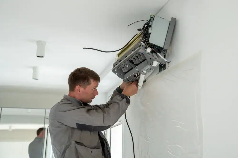 A technician in a gray uniform, from an HVAC Contractor Nassau County, NY, repairs or installs a wall-mounted air conditioning unit in a modern white room. Tools and exposed wires are visible as he works on the device.