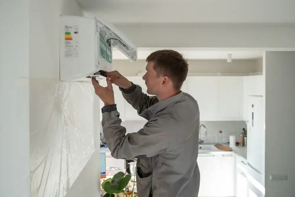 A technician in a gray uniform installs or repairs an air conditioning unit mounted on a wall inside a modern, white kitchen, showcasing the expertise of a professional HVAC Contractor in Nassau County, NY.