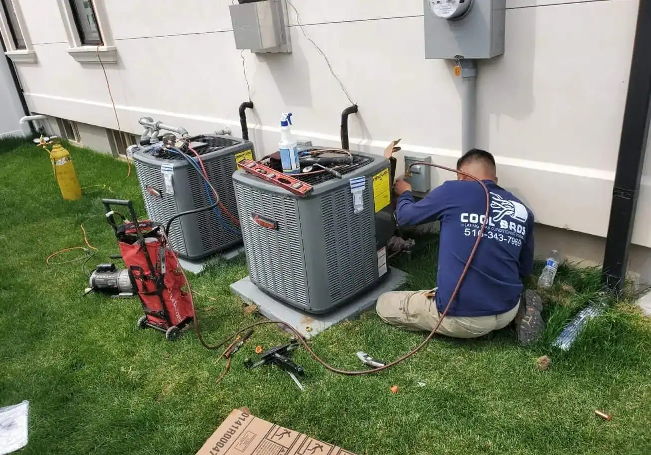 A technician in a "Cool Bros" shirt, representing an HVAC Contractor Nassau County, repairs an outdoor air conditioning unit next to a building in NY. Tools and equipment are scattered on the grass nearby, with two AC units and electrical meters visible.