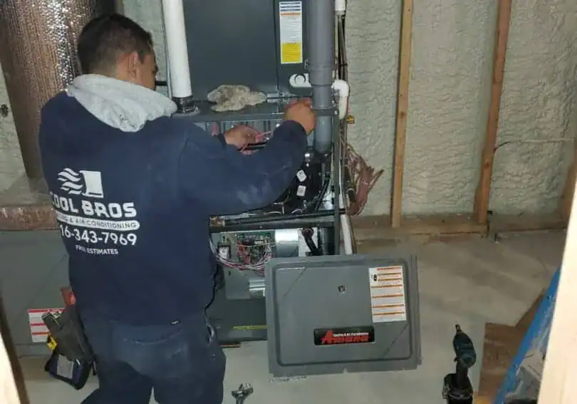 A technician in a “Cool Bros” sweatshirt, representing an expert HVAC Contractor in Nassau County, NY, repairs or installs a furnace in an unfinished basement. The open panel reveals wiring and components, with tools and a drill on the floor nearby.