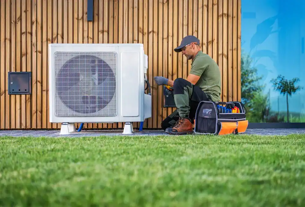 A technician from an HVAC Contractor in Nassau County, NY, wearing work clothes and a cap, repairs an outdoor air conditioning unit beside a modern wooden house. Tool bags rest nearby as he works on the unit while seated on the ground.
