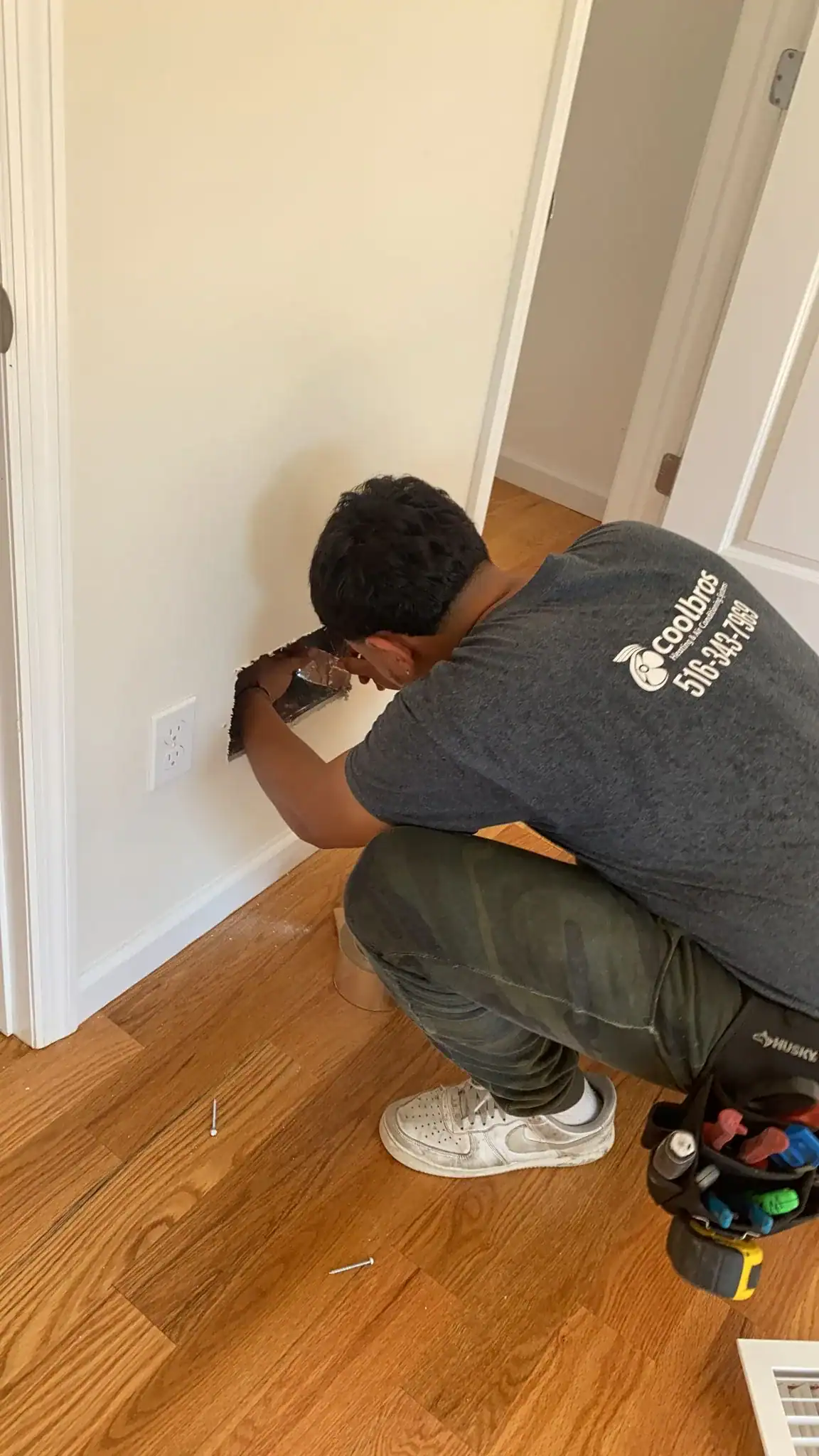 A person wearing a tool belt and a &ldquo;Goodzer S.O.G. Tech&rdquo; shirt kneels on a wooden floor, working on a large rectangular hole in a white wall near an electrical outlet&mdash;showcasing the expertise of an HVAC contractor in Nassau County, NY.