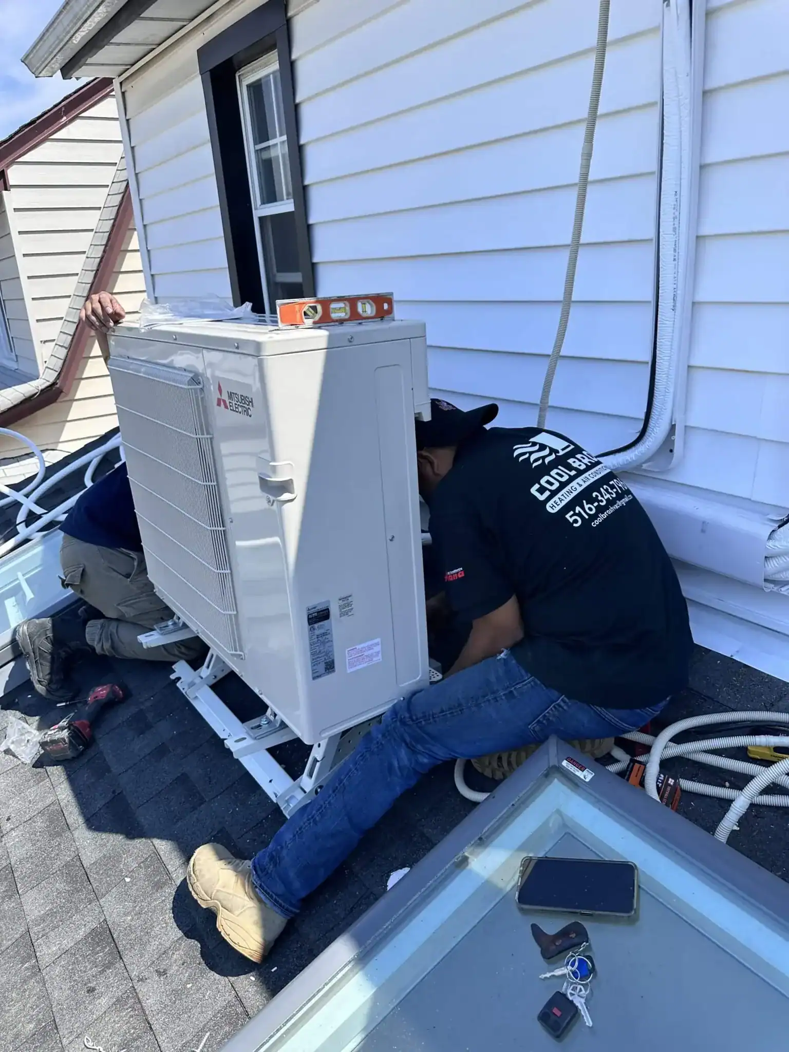 Two workers from an HVAC Contractor Nassau County install an outdoor air conditioning unit on a NY rooftop. Tools and cables are scattered around, and one worker in a black shirt and tan boots crouches beside the unit.